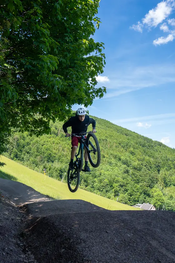 Biker přeskakuje terénní skok nad zelenou loukou v bikeparku Opálená